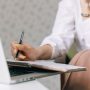 Closeup woman is holding notebook and making notes during online session, background internet laptop with web call.
